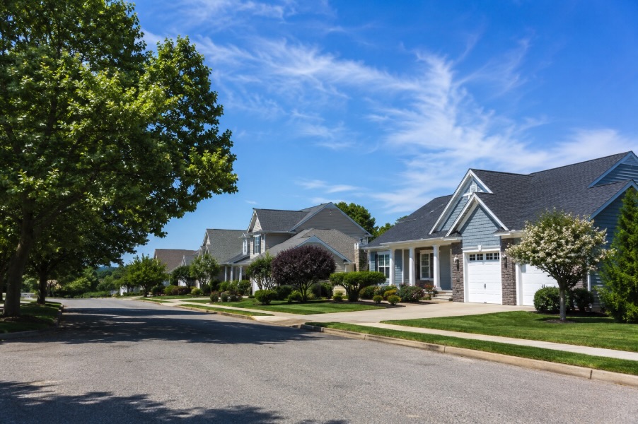 Wide landscape photo of a quiet residential neighborhood in coastal New Jersey on a clear sunny day, featuring a row of single-family homes with newly installed architectural shingle roofs, neatly maintained lawns, and mature trees lining the street beneath a bright blue sky with thin white clouds.