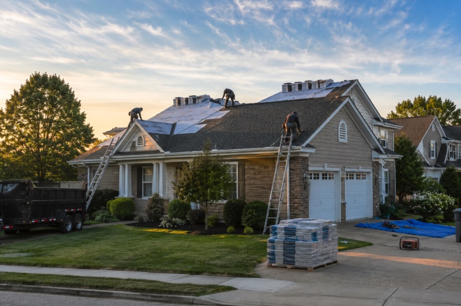 Wide landscape photo of a roofing crew working on a residential home in a quiet New Jersey suburb during golden hour, with bundles of shingles staged on the roof, ladders positioned along the house, and a clean organized job site visible from the street under a clear evening sky.