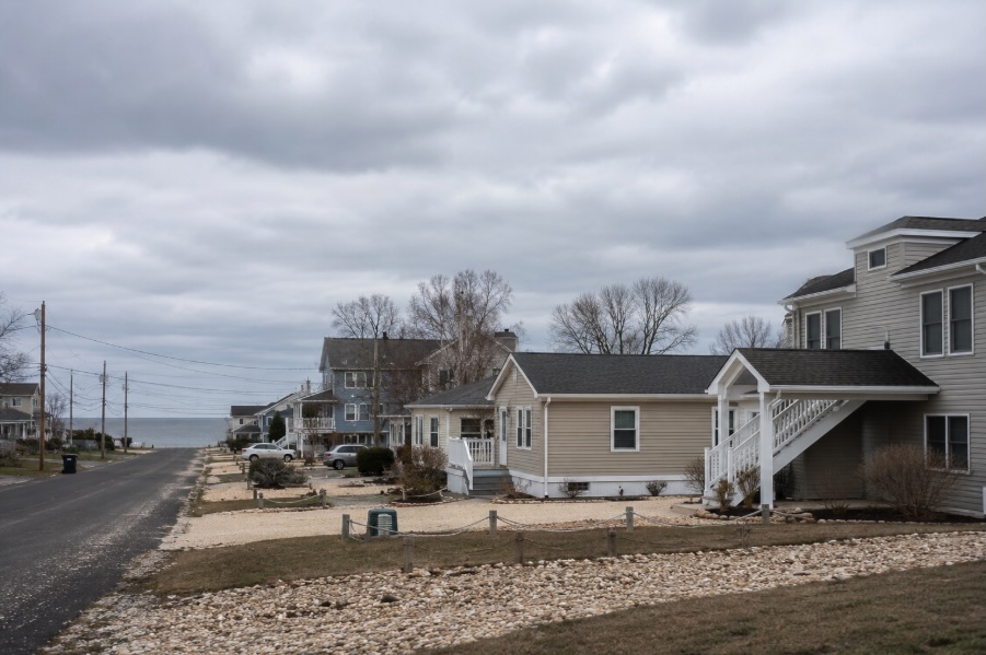 Wide view of a coastal New Jersey neighborhood with modest single-family homes, bare winter trees, and an overcast grey sky creating a cold freeze-thaw atmosphere.
